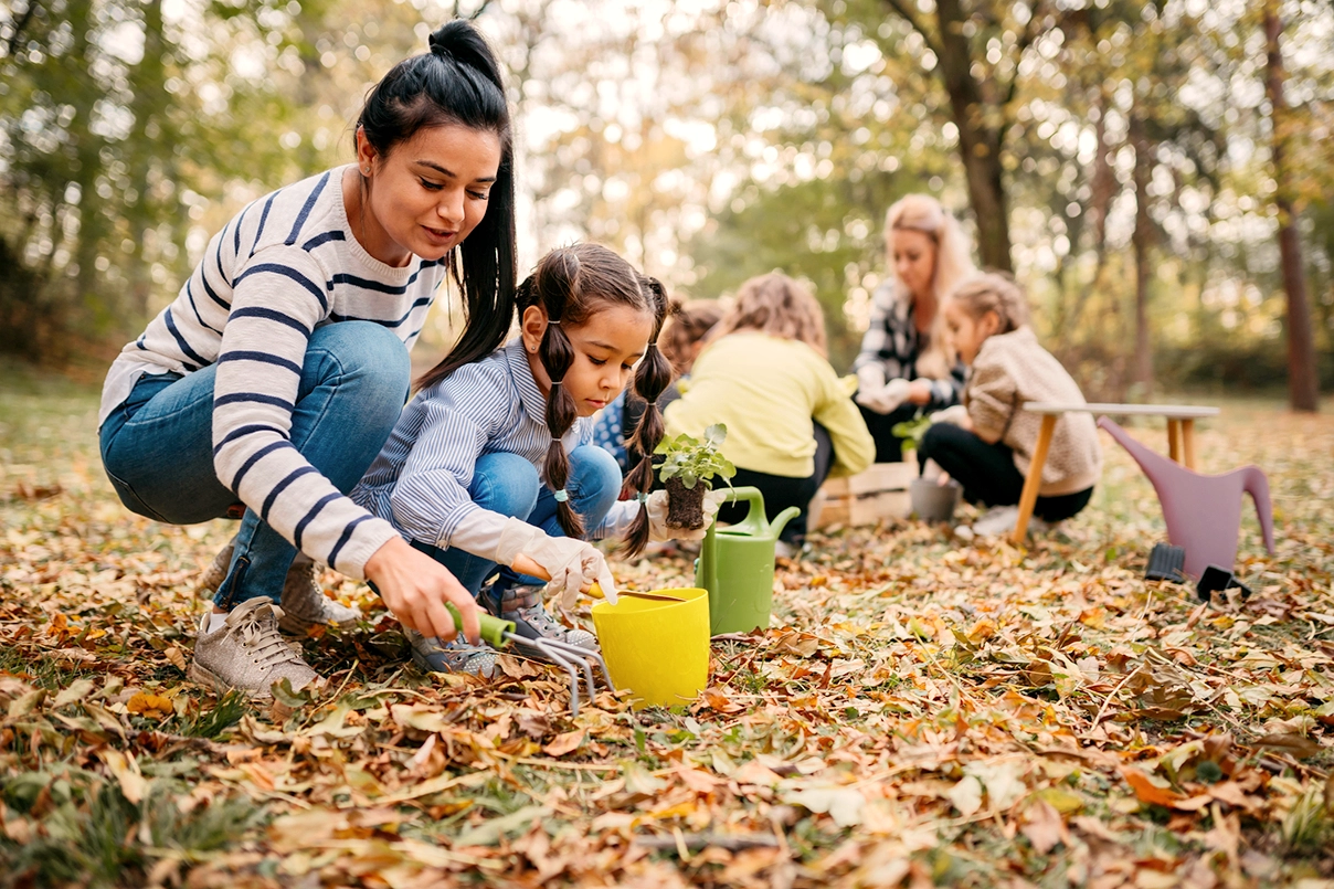 Zwei Frauen mit einer Gruppe von Kleinkindern mit Harken und Gießkanne im Wald. 